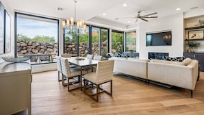 A modern living room with a dining area, large windows, a stone wall, and a ceiling fan. - Snow Canyon Sanctuary #8 4 Bedroom Home (Ivins)