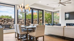 A modern living room with a stone wall, large windows, a dining area, and a ceiling fan. - Snow Canyon Sanctuary #8 4 Bedroom Home (Ivins)