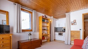 A room with a wooden ceiling, a TV on a dresser, a red sofa, a wooden bookshelf, a kitchen area with cabinets and a microwave, and a window with curtains. - 5m from ski slopes, floor -5, view ski slopes, balcony, 25m², Méribel-Mottaret (Méribel - Les Allues)