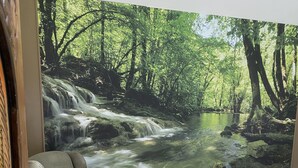A bedroom with a large forest-themed mural, a light-colored armchair, and a bed with a patterned bedspread. - New build OWL HOUSE, quiet, near trails, stargazing! (Landisburg)