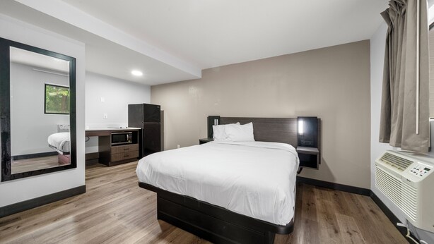 A modern hotel room with a large bed, a desk, a wardrobe, and a built-in air conditioning unit. - Southern Suites Carrollton (Carrollton)