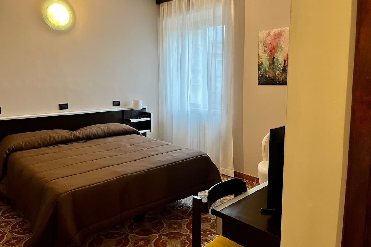 Ein Hotelzimmer mit Bett, Schreibtisch, Stuhl, einem Fenster mit Vorhängen und einem Gemälde an der Wand.
