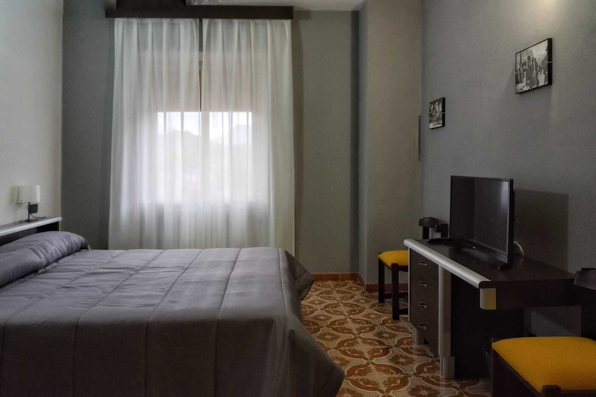 Ein Hotelzimmer mit einem Bett, einem Schreibtisch mit Fernseher, einem Sessel und einem kleinen Hocker.