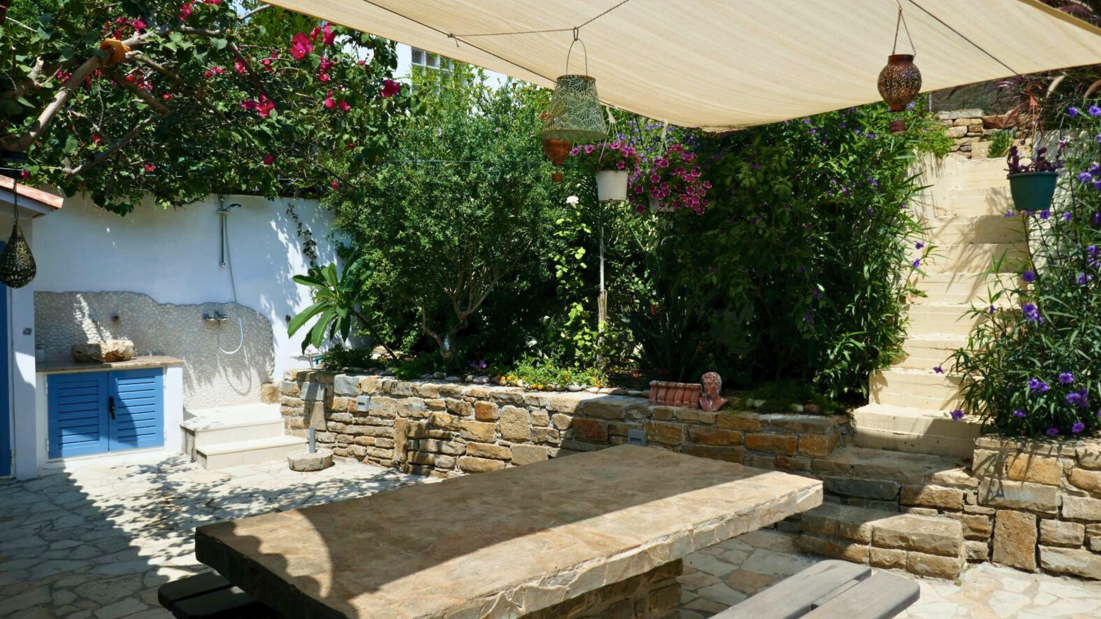 Um pátio externo com mesa e bancos de pedra, rodeado por vegetação exuberante e plantas floridas.