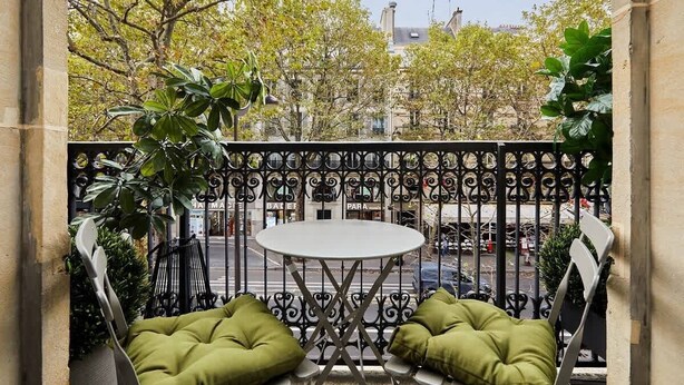 Ein Balkon mit rundem Tisch und zwei grünen, gepolsterten Stühlen, umgeben von einem schmiedeeisernen Geländer.