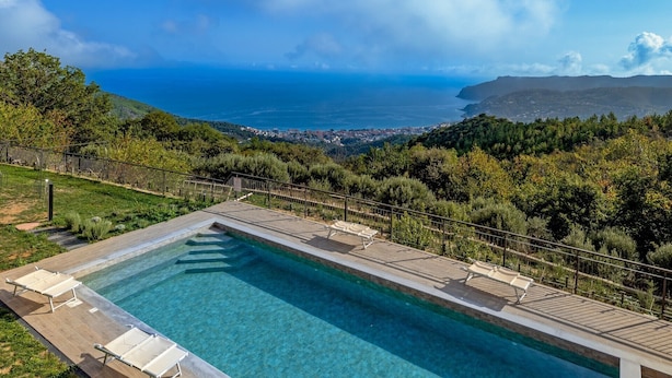 A rectangular pool with a wooden deck and lounge chairs, surrounded by a metal fence, overlooking a coastal landscape with trees and a distant town. - Agriturismo Cascina Trevo (Spotorno)