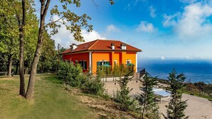 A two-story house with a red roof and orange walls, surrounded by greenery and overlooking a body of water. - Agriturismo Cascina Trevo (Spotorno)