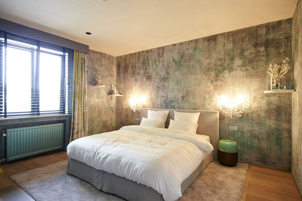 A bedroom with a large bed, a window with blinds, a radiator, and a green waste bin.