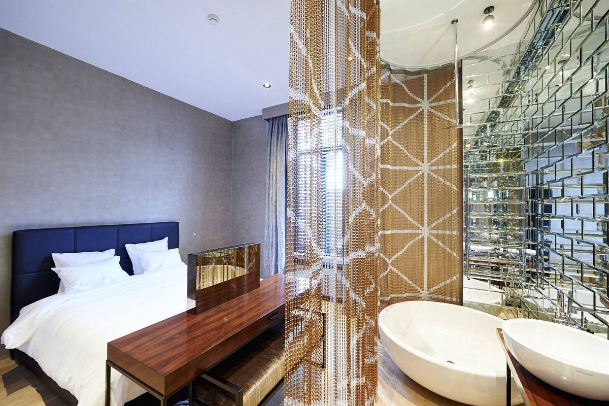 A modern hotel room with a large bed, a wooden side table, a large mirror, and a bathroom with a large bathtub and a glass-enclosed shower area.