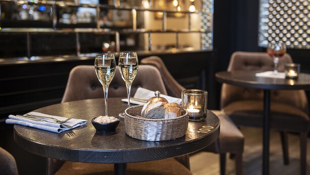 Een restauranttafel met een rond tafelkleed, een broodmand, twee glazen witte wijn, een kaars en een servet met bestek.