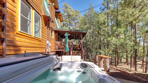 A hot tub in front of a wooden cabin with a porch and a view of pine trees. - Hot Tub + Decks: Sherwood Forest Cabin (Lakeside)