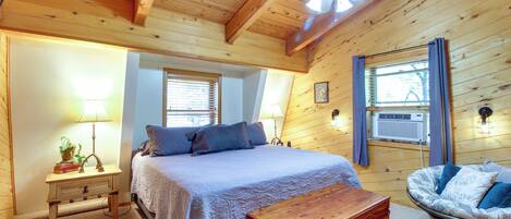 Un dormitorio de cabaña de madera con cama, mesita de noche, lámpara y sillón mecedor.