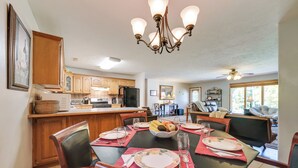 A dining area with a large table set for six, surrounded by wooden chairs. A chandelier hangs above, and a kitchen is visible in the background.