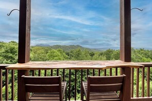 Terraza de madera con dos sillas y una mesa, con vistas a un paisaje boscoso y colinas.
