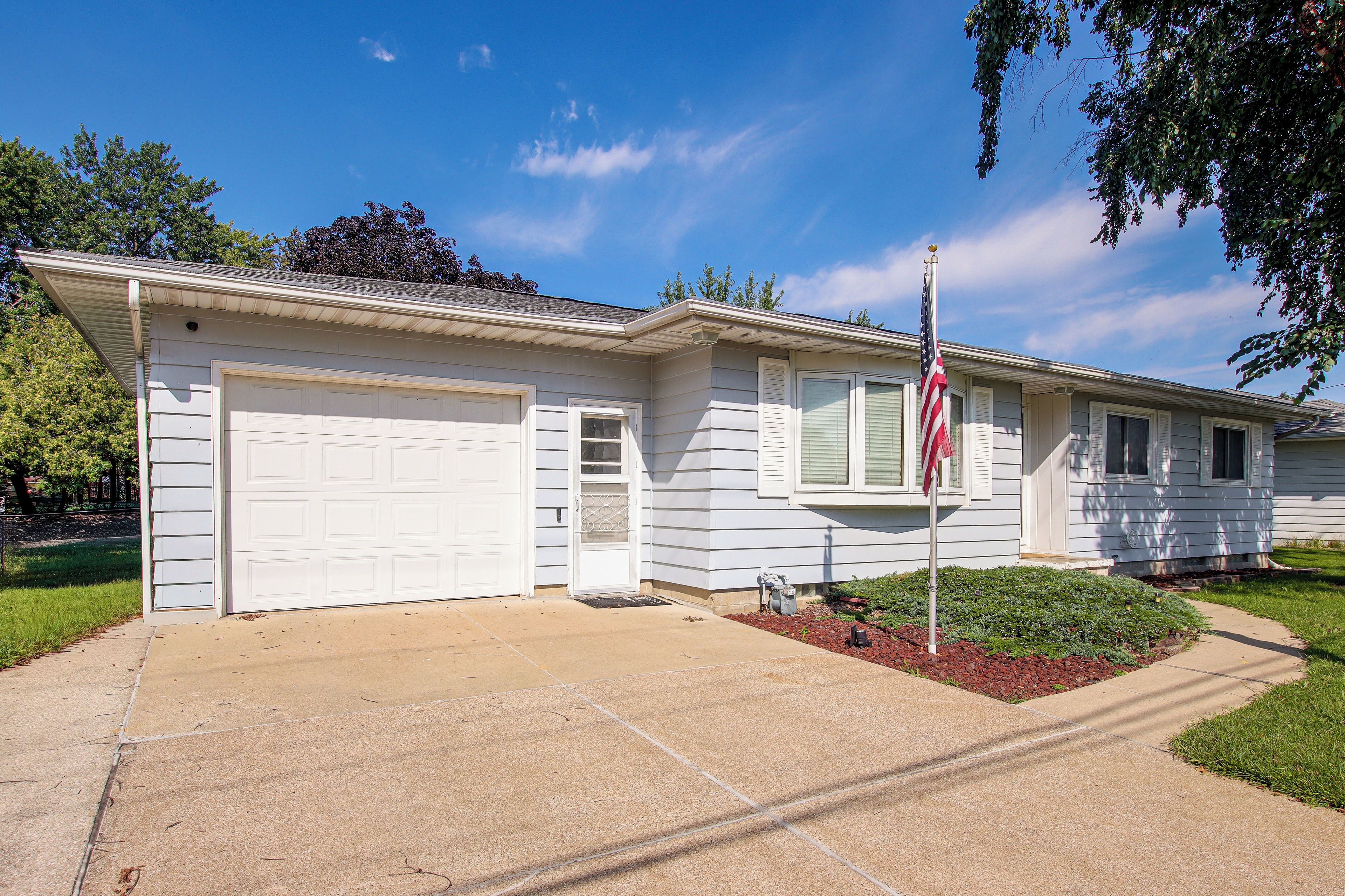 A single-story house with a garage, a front door, and a small front yard with a flagpole.
