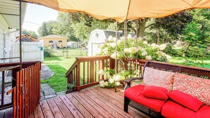 Une terrasse extérieure couverte, dotée d’une rambarde en bois, d’un banc rouge avec coussin et offrant une vue sur une cour arrière avec une maison mobile blanche et des arbres.