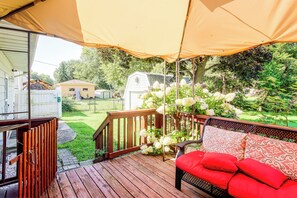 A covered outdoor deck with a wooden railing, a red cushioned bench, and a view of a backyard with a white mobile home and trees. - Pet-friendly Waterloo Abode w/ Deck! (Waterloo)