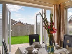 A dining area with a table set for two, a vase with flowers, and a view of a backyard through open doors. - 3 bedroom accommodation in Bridlington (Bridlington)
