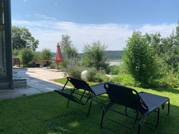A patio with two black lounge chairs, a red umbrella, and a view of a lake surrounded by greenery. - Stille x Weite - Hochwertige Wohnung mit See-panorama (Kronburg)