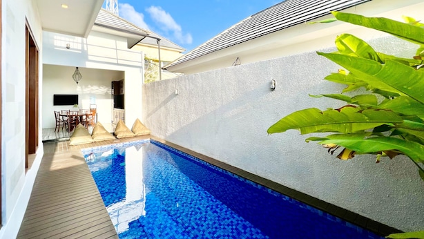 Un área de piscina moderna con una piscina de color azul cristalino, una terraza de madera y una pared blanca con textura.