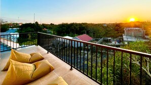 Un balcón con vistas a un atardecer, una piscina y un edificio lejano.