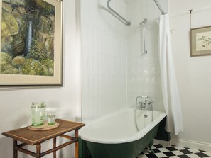 Un baño con una bañera tipo pata de león verde, paredes de azulejos blancos, un cuadro enmarcado y un suelo a cuadros.