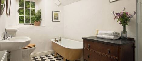 Un baño con piso a cuadros, una bañera, un lavabo, una tocador y una ventana con una planta.