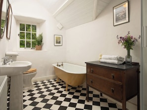 Un baño con piso a cuadros, una bañera, un lavabo, una tocador y una ventana con una planta.
