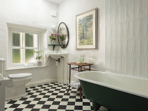 Un baño con piso a cuadros, una bañera verde tipo pata de león, un inodoro blanco, un lavamanos con espejo y un cuadro en la pared.