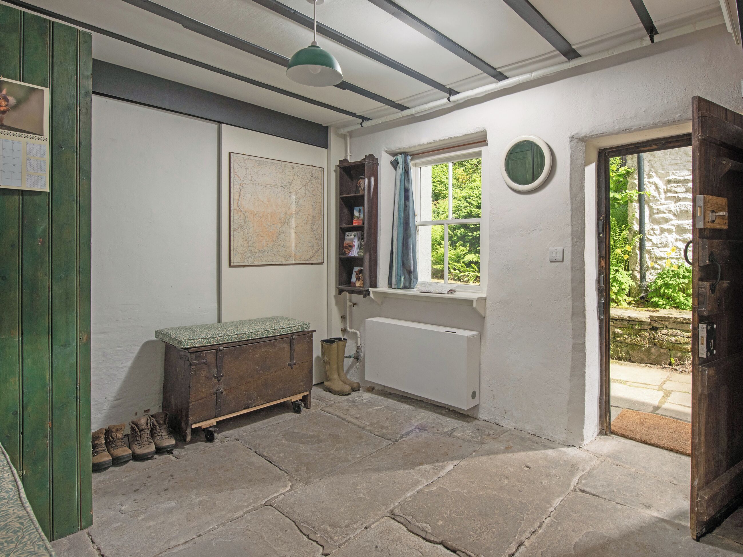 A room with a radiator, a window with a view of greenery, a wooden cabinet with boots, and a map on the wall.