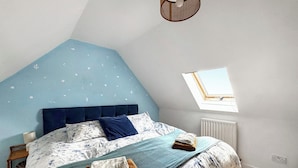 Une chambre avec un plafond en pente, un lit recouvert d’une literie à motifs floraux, une fenêtre et un radiateur.