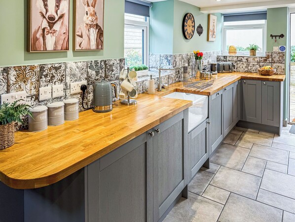 A kitchen with a wooden countertop, grey cabinets, a kettle, and a clock on the wall.