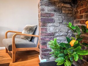 Une chaise en bois avec un coussin gris et un coussin décoratif, placée à côté d’un mur de briques, devant une plante aux feuilles vertes et un fruit orange.