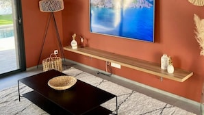 Ein modernes Wohnzimmer mit einem großen Flachbildfernseher an der Wand, einem dunklen Couchtisch, einem weißen Teppich mit Struktur und Holzregalen mit Dekorationsobjekten.