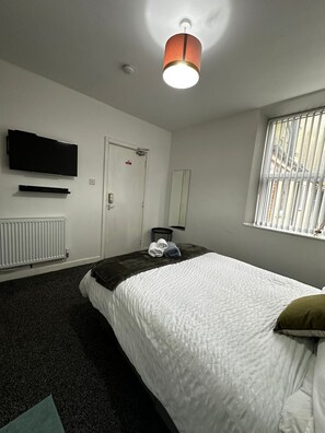 A hotel room with a bed, a television, a radiator, a door, and a window with blinds. - Blackwood Residence (Accrington)