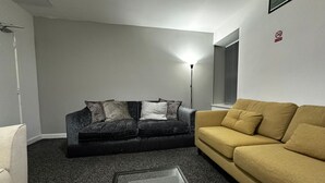 A living room with a dark grey sofa, a yellow sofa, and a white armchair. There is a floor lamp and a window with blinds. - Blackwood Residence (Accrington)