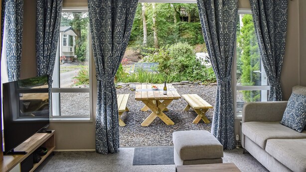 Un salon chaleureux avec une télévision, un canapé et une table en bois avec des bancs. De grandes fenêtres ornées de rideaux à motifs offrent une vue sur un jardin et une maison.