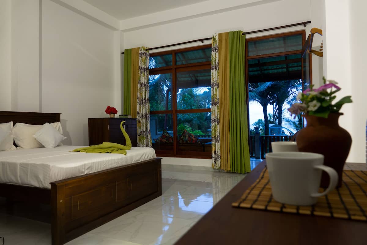 A hotel room with a bed, a window with curtains, a vase with flowers, and a table with a cup.