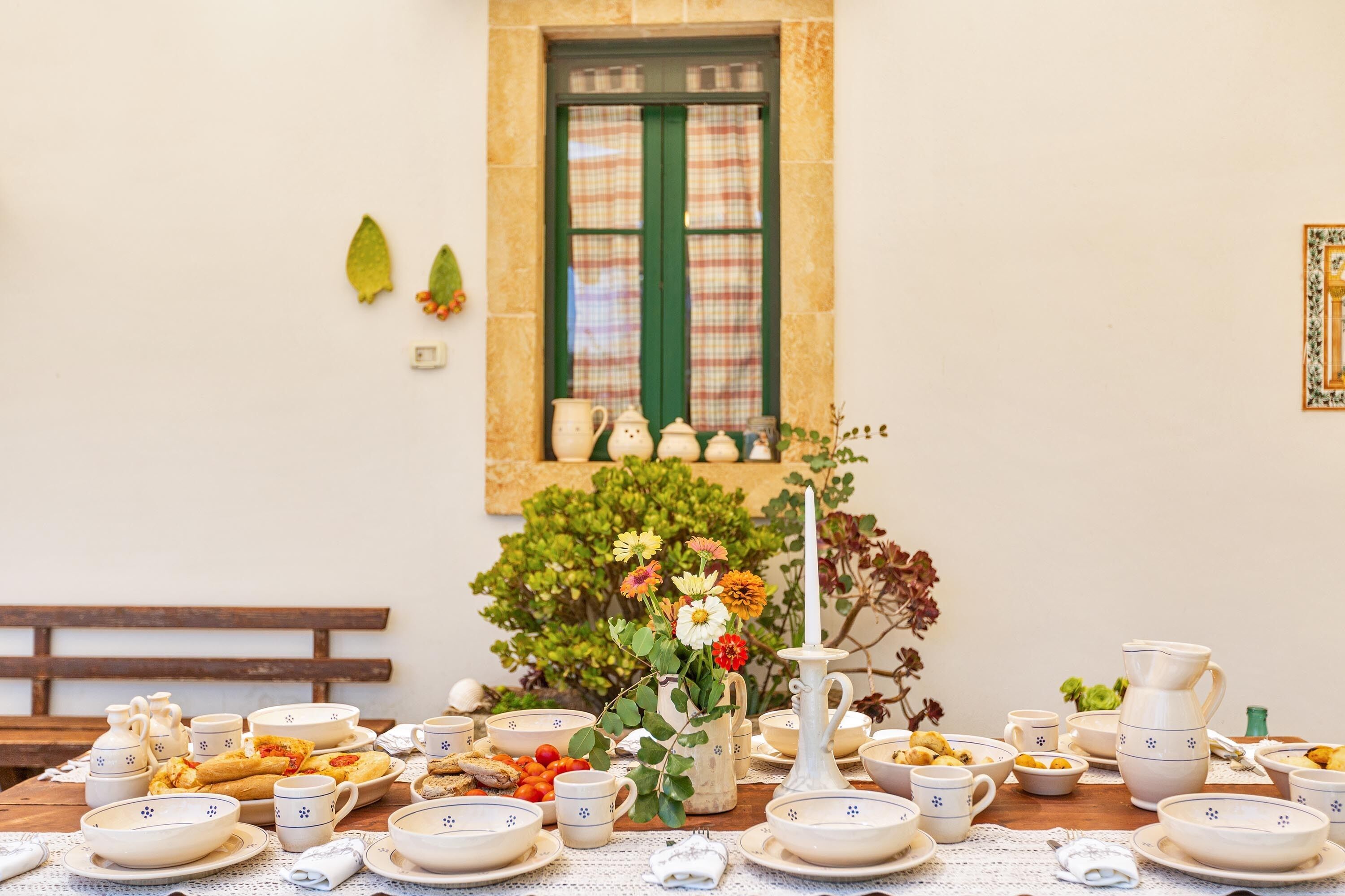 Une table à manger dressée avec de la vaisselle blanche et bleue à motifs, un vase rempli de fleurs et une fenêtre verte ornée de rideaux à carreaux.