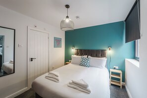 A neatly made bed with towels, a bedside table, a nightstand, a wall-mounted TV, and a door with a mirror. - Deluxe Studio Apartments (Derby)