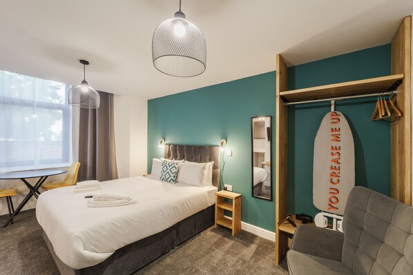 A modern hotel room with a large bed, a desk, a chair, a wardrobe, and a large window. - Deluxe Studio Apartments (Derby)