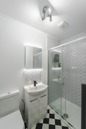A modern bathroom with a glass shower enclosure, a white sink, and a herringbone-patterned tile backsplash. - Deluxe Studio Apartments (Derby)