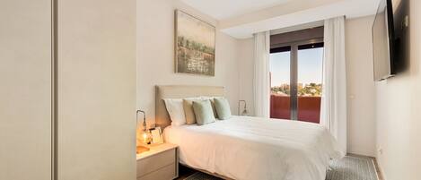 A modern bedroom with a large bed, a nightstand, a painting, a TV, and a view of the outdoors through a sliding door.