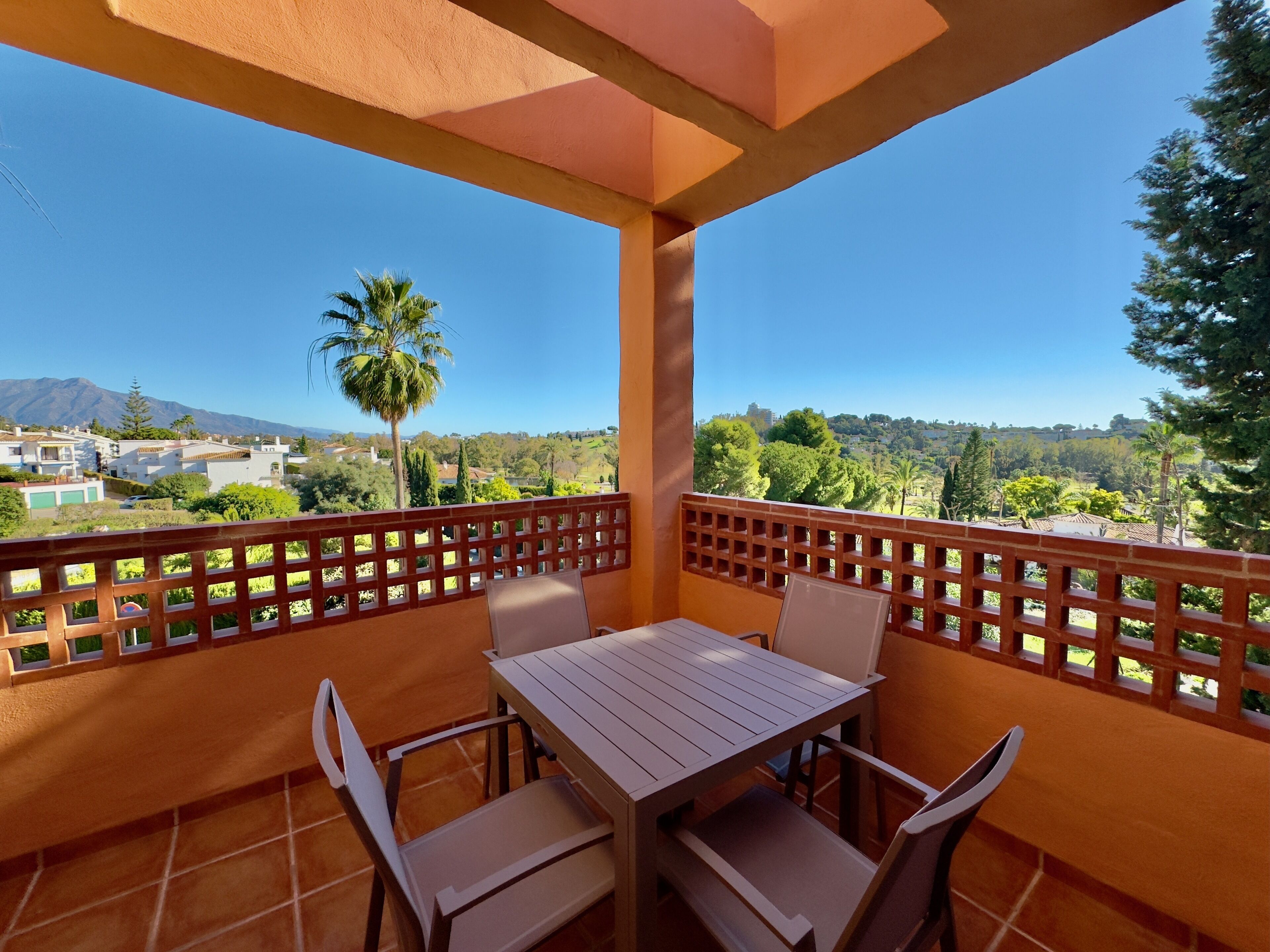 A balcony with a table and chairs, offering a view of a residential area with trees and mountains in the distance.