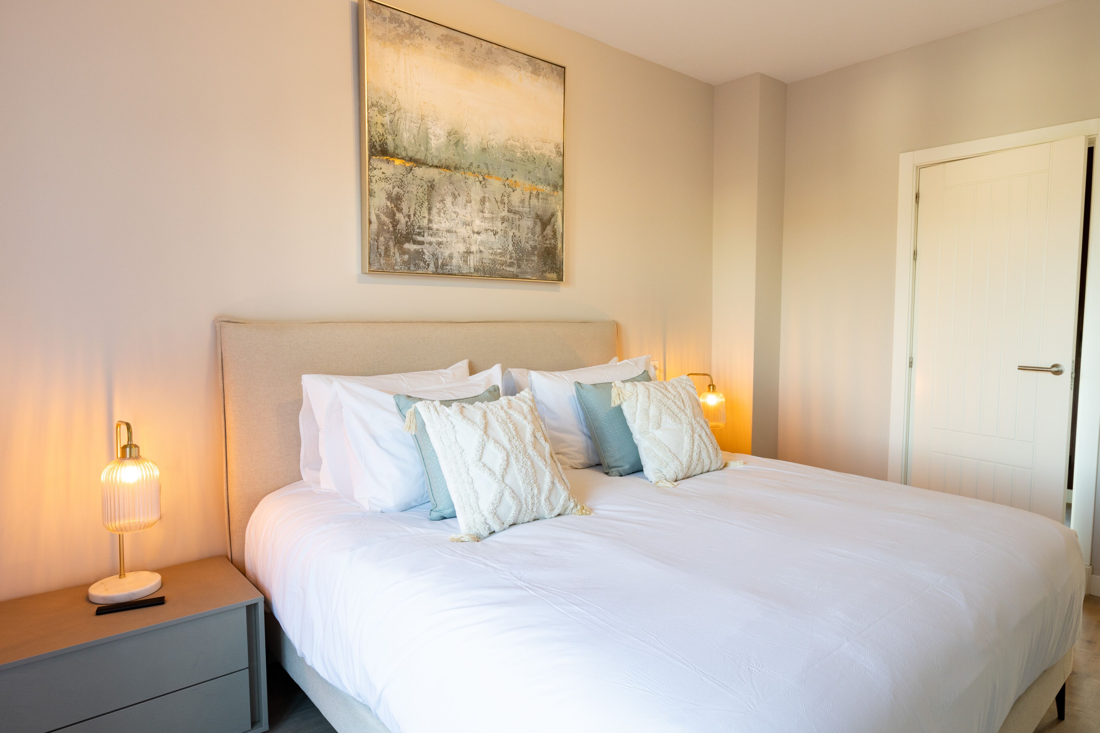 A neatly made bed with white linens and several pillows, a bedside table with a lamp, and a painting on the wall.