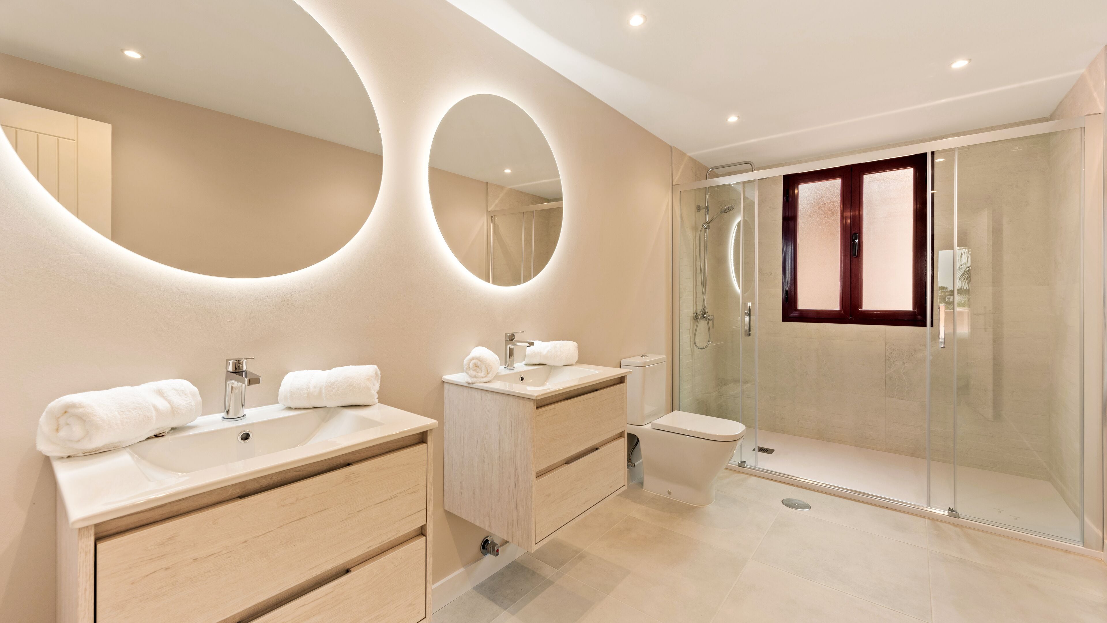 Un baño moderno con dos lavamanos, cabina de ducha con mampara de vidrio y un espejo grande con iluminación circular.