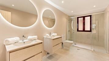 Un baño moderno con dos lavamanos, cabina de ducha con mampara de vidrio y un espejo grande con iluminación circular.