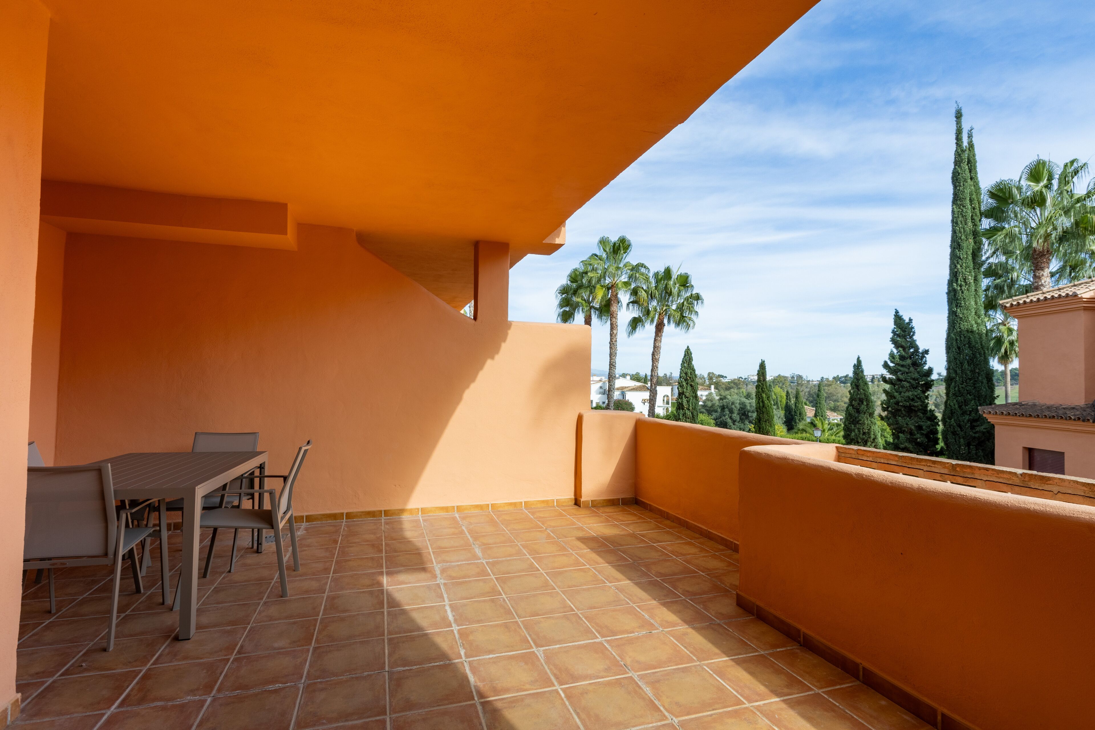 A terrace with a table and chairs, a tiled floor, and a view of palm trees and buildings.