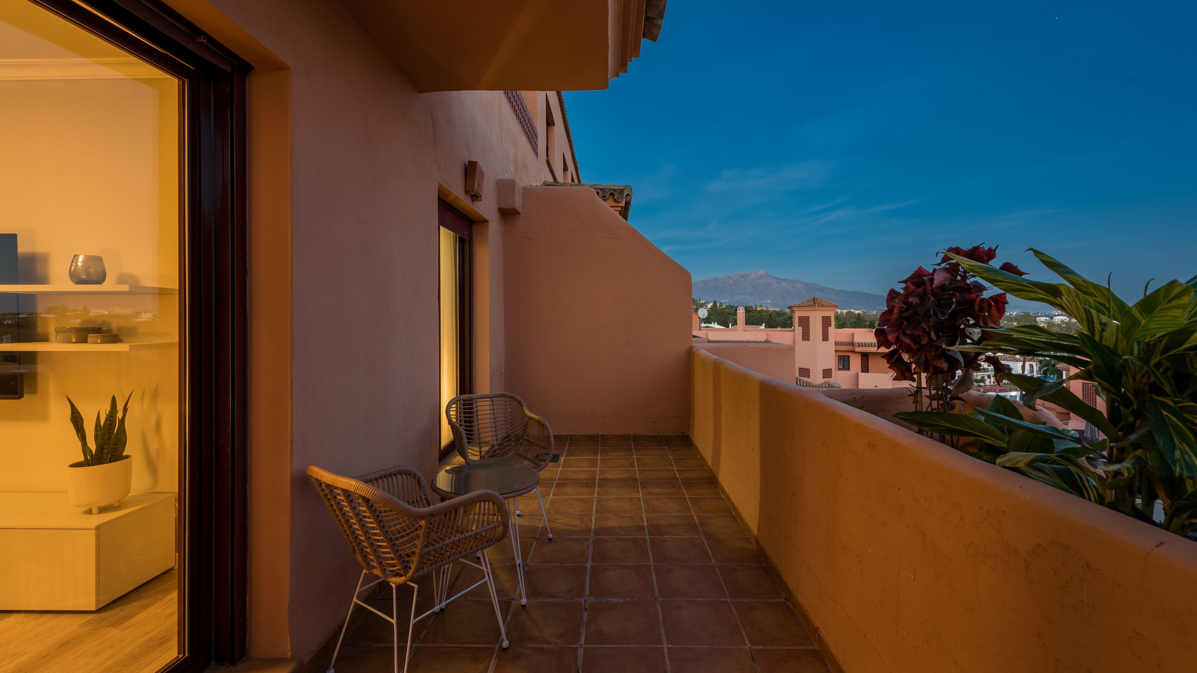 A balcony with a table and chairs, a potted plant, and a view of a mountain in the background.