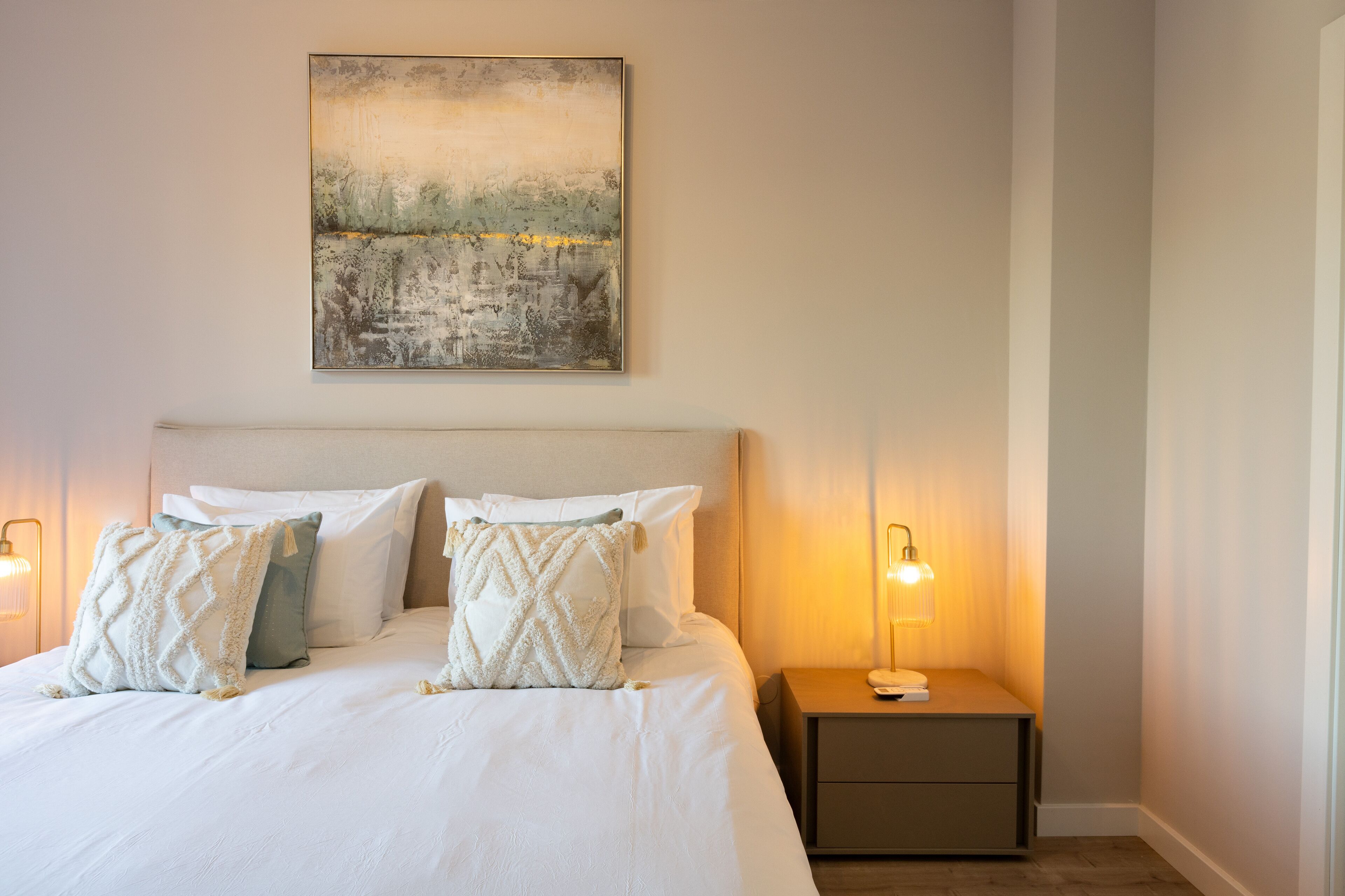 A neatly made bed with white linens and decorative pillows, a bedside table with a lamp, and a painting on the wall.